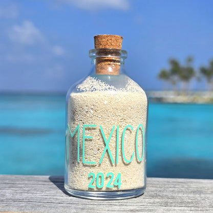 Glass bottle with cork lid containing sand labeled 'Mexico 2024' against a beach background