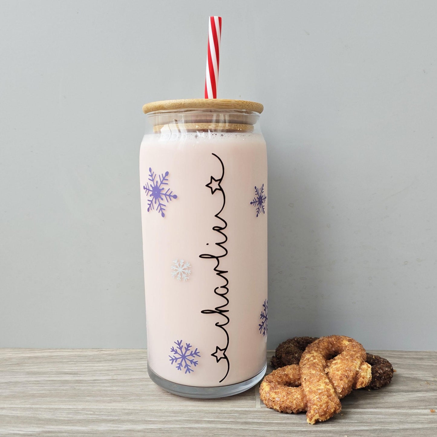 Personalised glass can drink holder with a bamboo suction lid and red and white reusable straw. Features silver snowflakes and blue snowflakes that colour change to purple when cold liquid is added to the glass. Personalised name along the side of the glass, available in glittery silver or black and features either a heart or stars at either end of the name.