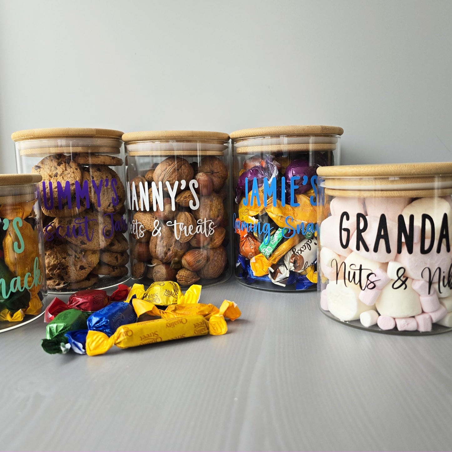 Four glass jars with wooden lids, each labeled with a different name, filled with various snacks on a gray surface.